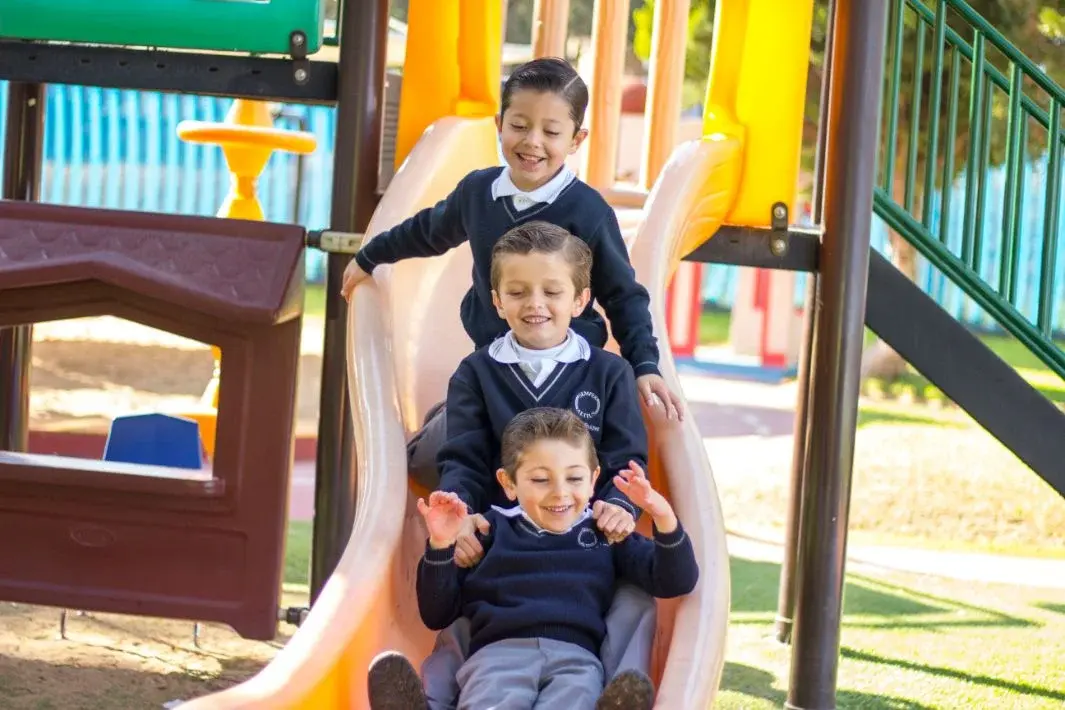 Tres estudiantes de uniforme escolar disfrutando juntos de un tobogán en el patio de recreo del colegio.
