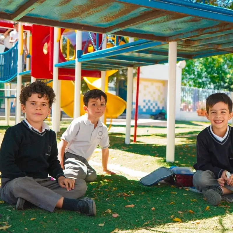 Tres estudiantes sentados en el área de juegos del colegio durante recreo, bajo estructura de sombra colorida.